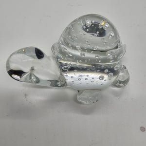 Puede incluir: Una figura de tortuga de cristal transparente con caparazón redondeado y burbujas visibles. La tortuga tiene un cuerpo y cabeza lisos y transparentes, con pequeñas aletas redondeadas. Es un artículo decorativo.