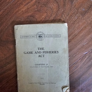 May include: A vintage book titled "The Game and Fisheries Act" from the Department of Mines and Natural Resources. The cover is aged, with text including "Chapter 15, Statutes of Manitoba, 1930" and the year 1932. The book is on a wooden surface.