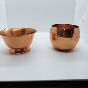 Può includere: Due ciotole color rame. Una è svasata con base a piedistallo, l'altra è di forma rotonda e sferica. Entrambe hanno una superficie lucida e riflettente, su sfondo bianco.