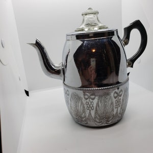 Puede incluir: Una cafetera cromada vintage con tapa de cristal y asa negra. La parte inferior de la cafetera presenta un diseño floral grabado. El pico es curvo y la superficie es brillante y reflectante.