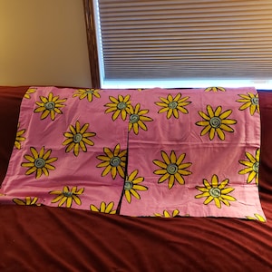 Puede incluir: Fundas de almohada rosas con un patrón repetido de margaritas amarillas con contornos negros y centros azules. Las fundas de almohada están dobladas sobre una superficie roja oscura. Las margaritas están estilizadas y añaden un toque alegre a la ropa de cama.