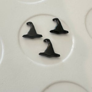 May include: Three black witch hats on a white surface. The hats are small and made of a shiny material.