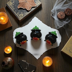 May include: Three black cauldron-shaped decorations with green bubbling potion and flames. The cauldrons sit on a white paper with candles, books, and other Halloween-themed items. The scene is set on a dark wooden surface.