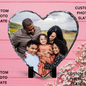 Op de afbeelding: Een hartvormig fotolijstje van leisteen met een familiefoto van een man, een vrouw en drie kinderen. Het lijstje staat op een roze houten oppervlak met witte bloemen.