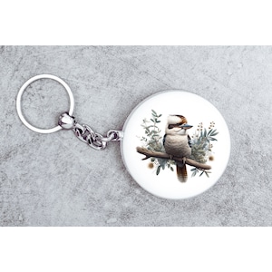 May include: A silver key chain with a round, white charm featuring a detailed illustration of a kookaburra perched on a branch with green foliage.