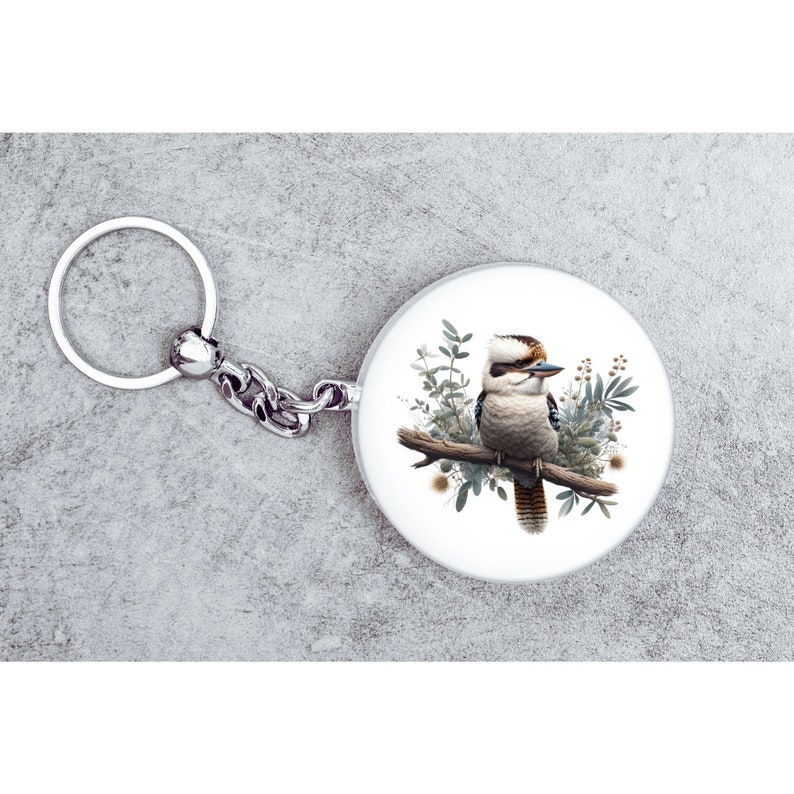 May include: A silver key chain with a round, white charm featuring a detailed illustration of a kookaburra perched on a branch with green foliage.