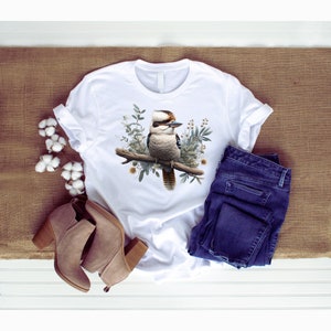 May include: White t-shirt featuring a kookaburra bird perched on a branch with green leaves. The bird is brown, white and black with a long tail.