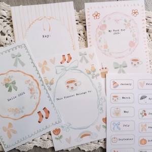Puede incluir: Una colección de artículos de papelería, incluyendo páginas de planificador y pegatinas, con un diseño de acuarela suave. Las páginas presentan ilustraciones florales y lazos, con texto que dice "Hello 2026" y "My Word for 2026". Las pegatinas están etiquetadas con meses.
