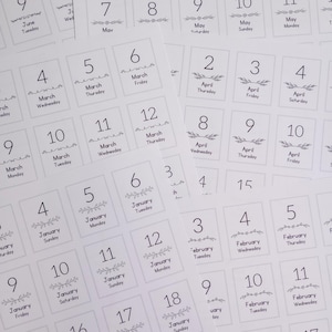 May include: A collection of white square cards with dates and days of the week printed in black. Each card features a number, month, and day, with decorative floral elements. The cards are scattered across a white surface.