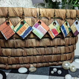 May include: A collection of six diamond-shaped earrings with colorful, patterned paper inserts. The earrings hang from bronze-colored hooks and are displayed against a woven basket background. The paper designs feature a collage of colors and textures.