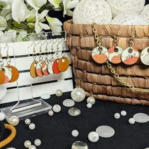 May include: A display of handmade earrings with autumn-themed designs. The earrings feature pumpkin and leaf motifs in orange, white, and brown. Some earrings are displayed on a clear stand, while others are in a woven basket. Decorative pearls and floral accents complete the scene.