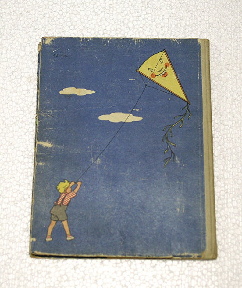 May include: A vintage children's book cover with a blue background and a child flying a kite. The kite is yellow with a smiling face and red cheeks. The text on the cover reads "23 июл."