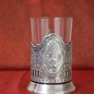 May include: Silver-plated glass holder with an ornate design featuring a bird and floral motifs. The holder is designed to hold a small glass or cup.