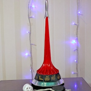 以下が含まれることがあります： 赤と銀色のロケット型のランプ。黒いベースと白い電源コードが付いています。 ランプには、金色のアクセントが付いた詳細なベースがあります。 背景には青いライトのストリングがあります。