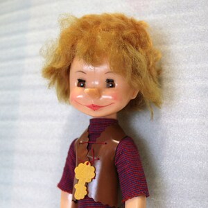 May include: A vintage doll with a cheerful expression, featuring a reddish-brown vest over a striped shirt. The doll has orange hair and a yellow key-shaped pendant. The doll's face is painted with rosy cheeks and a red smile. A collectible toy.