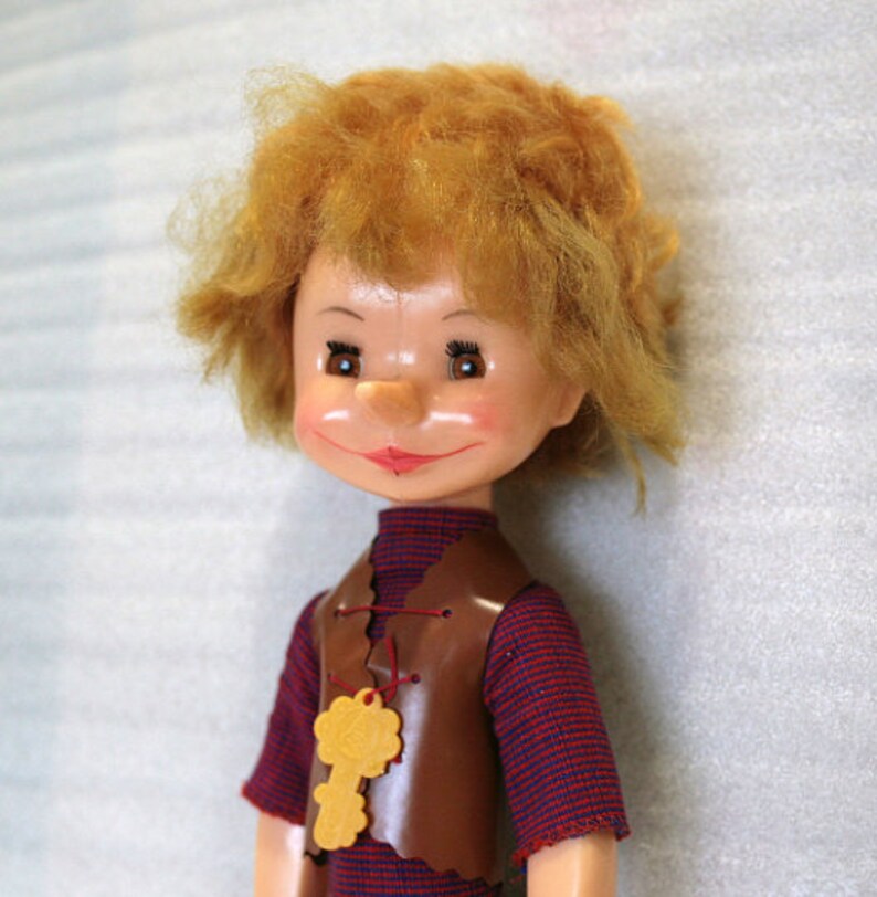 May include: A vintage doll with a cheerful expression, featuring a reddish-brown vest over a striped shirt. The doll has orange hair and a yellow key-shaped pendant. The doll's face is painted with rosy cheeks and a red smile. A collectible toy.