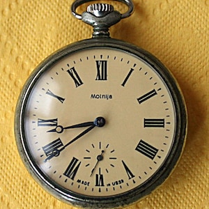May include: A silver pocket watch with Roman numeral markings on the face. The watch face is white with black hands and the word "Molnija" printed in black. The watch is made in the USSR.