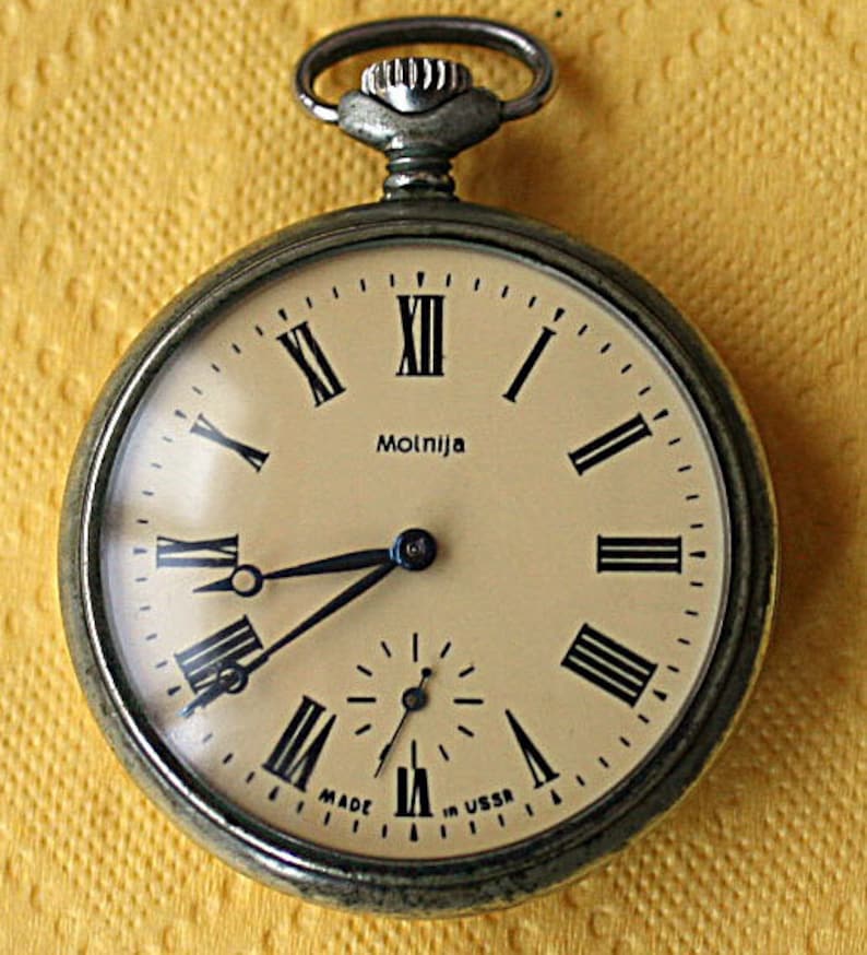 May include: A silver pocket watch with Roman numeral markings on the face. The watch face is white with black hands and the word "Molnija" printed in black. The watch is made in the USSR.