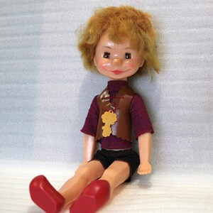 May include: A vintage doll with a painted face, red hair, and a brown vest with a key-shaped pendant. The doll wears a red and blue striped shirt, black shorts, and red shoes. The doll is sitting down.