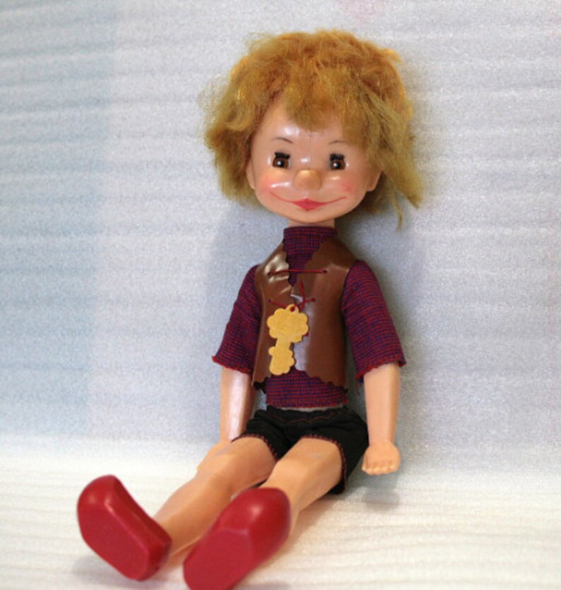 May include: A vintage doll with a painted face, red hair, and a brown vest with a key-shaped pendant. The doll wears a red and blue striped shirt, black shorts, and red shoes. The doll is sitting down.