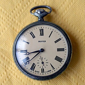 May include: A silver pocket watch with Roman numeral markings on the face. The watch face is cream colored with black markings and the word "Molnija" printed on it. The watch has a silver case and a silver chain attached to the top.