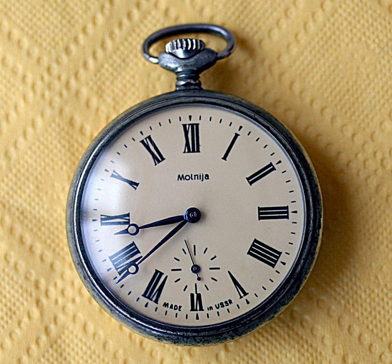 May include: A silver pocket watch with Roman numeral markings on the face. The watch face is cream colored with black markings and the word "Molnija" printed on it. The watch has a silver case and a silver chain attached to the top.