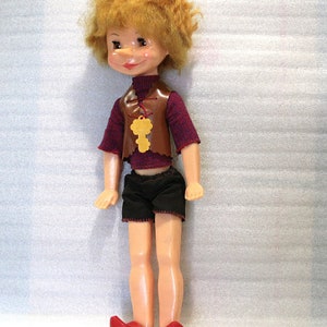 May include: A vintage doll with a painted face, red hair, and a brown vest with a key pendant. The doll wears a maroon shirt, black shorts, and red shoes. The doll is standing upright.
