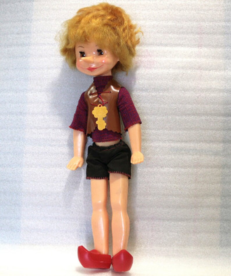 May include: A vintage doll with a painted face, red hair, and a brown vest with a key pendant. The doll wears a maroon shirt, black shorts, and red shoes. The doll is standing upright.