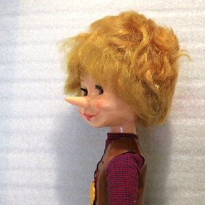 May include: A vintage doll with a long nose, blonde hair, and a smiling expression. The doll wears a brown vest over a red and purple patterned shirt. A gold-colored medallion hangs from the vest.