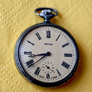 May include: A silver pocket watch with Roman numeral markings on the face. The watch face is cream colored with black hands and a black second hand. The watch face also has the text "Molnija" and "Made in USSR".