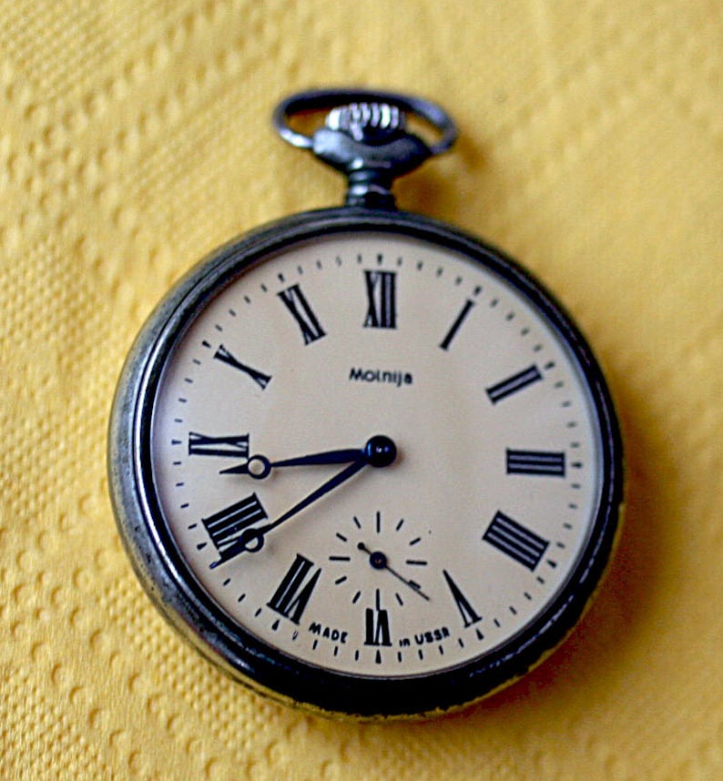 May include: A silver pocket watch with Roman numeral markings on the face. The watch face is cream colored with black hands and a black second hand. The watch face also has the text "Molnija" and "Made in USSR".