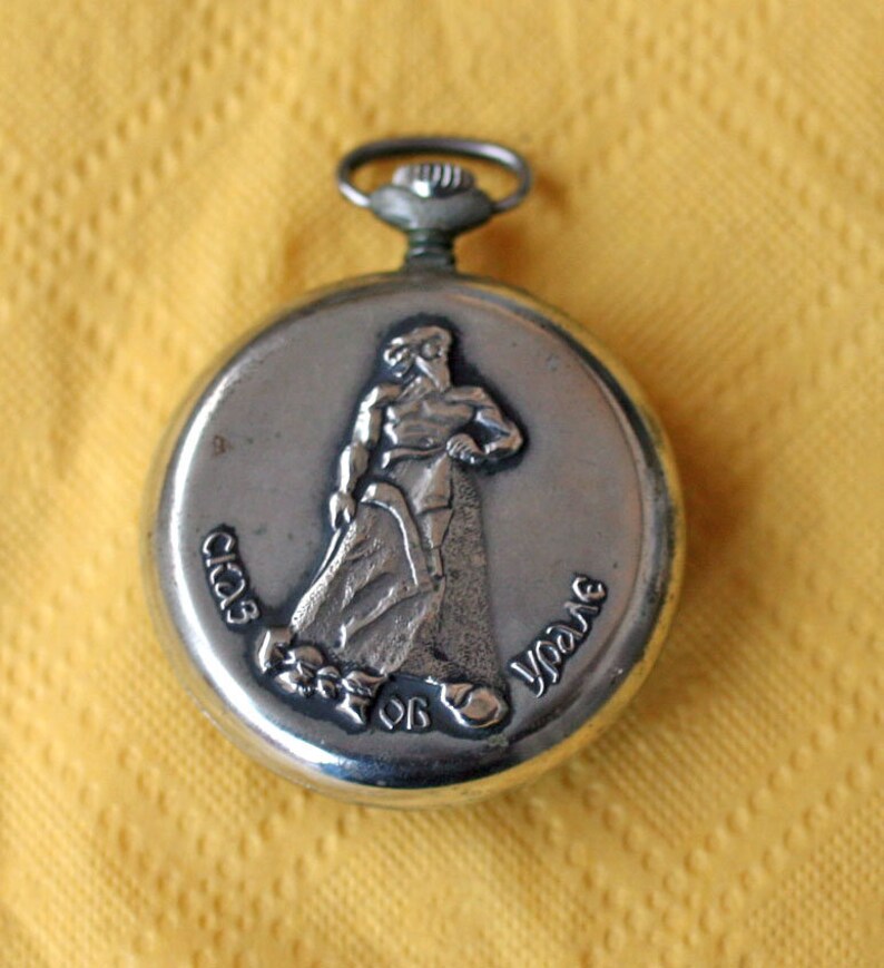 May include: Silver pocket watch with a raised image of a person holding a rope. The watch face reads "Club de Yo-Yo".