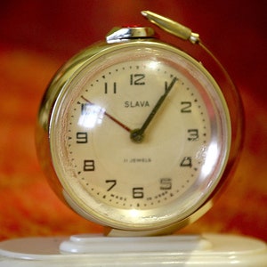Può includere: Un orologio da sveglia vintage color oro con quadrante bianco e lancette nere. Il quadrante dell'orologio ha numeri romani neri per le ore e indici neri per i minuti. Il quadrante dell'orologio riporta anche la scritta "SLAVA" e "15 JEWELS".