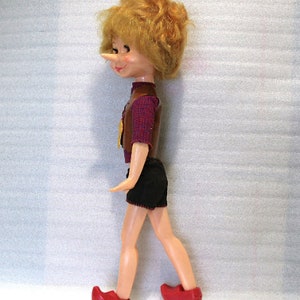 May include: A vintage doll of a boy with a long nose, wearing a brown vest over a red plaid shirt, black shorts, and red shoes. The doll has blonde hair and is in a walking pose.