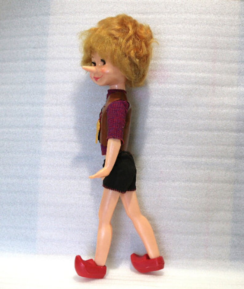 May include: A vintage doll of a boy with a long nose, wearing a brown vest over a red plaid shirt, black shorts, and red shoes. The doll has blonde hair and is in a walking pose.
