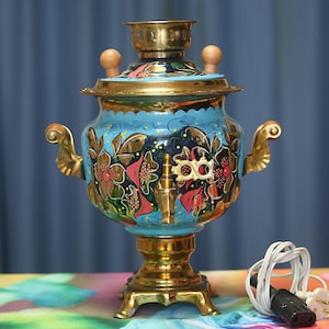 May include: A bright blue electric samovar featuring a hand-painted floral design with gold, red, and dark green accents. The metal vessel has a polished brass base, lid, and side handles, with two wooden knobs on top and a white power cord attached.