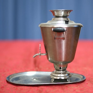 May include: A silver samovar with a black handle and a spout on a silver tray. The samovar is a traditional Russian tea kettle.