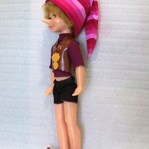 May include: A vintage doll dressed as a storybook character. The doll has a long nose, blonde hair, and is wearing a striped pink and red hat, a brown vest, black shorts, and red shoes. A gold-colored key hangs from the vest.