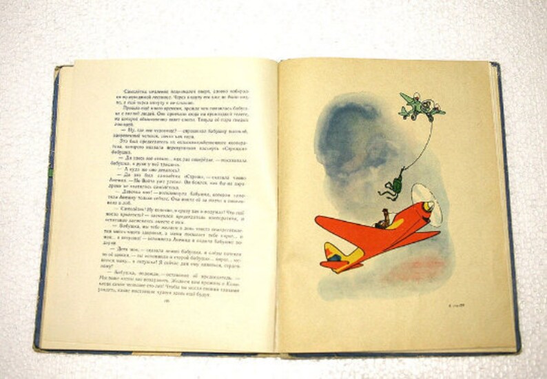 May include: An open book with a red and yellow airplane illustration on the right page. The left page has text in Russian. The book is on a white surface.