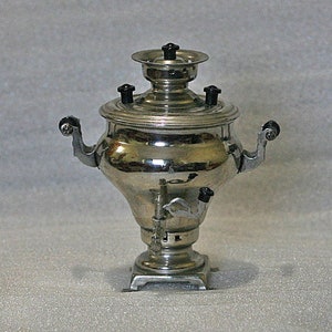 May include: A silver samovar with black accents and a spout on the side. The samovar has a round body and a square base.