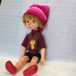 May include: A vintage doll with blonde hair, rosy cheeks, and a whimsical expression. It wears a striped pink and red hat, a brown vest with a key pendant, a red and blue striped shirt, black shorts, and red shoes. The doll is sitting.