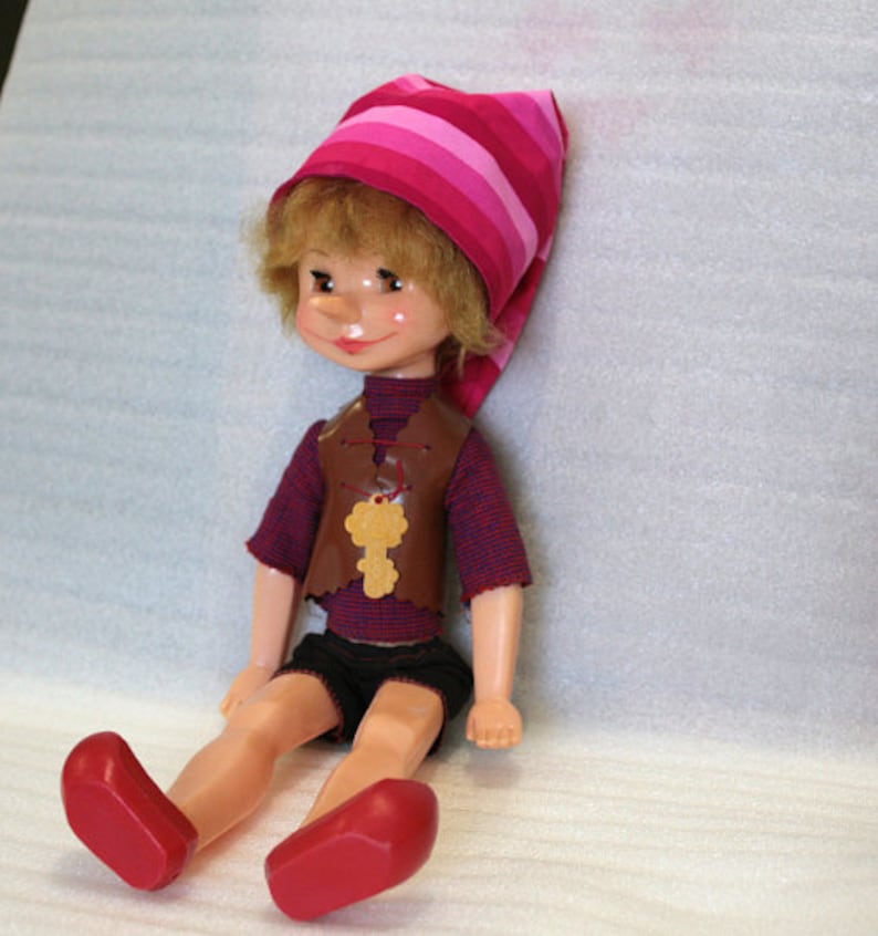 May include: A vintage doll with blonde hair, rosy cheeks, and a whimsical expression. It wears a striped pink and red hat, a brown vest with a key pendant, a red and blue striped shirt, black shorts, and red shoes. The doll is sitting.