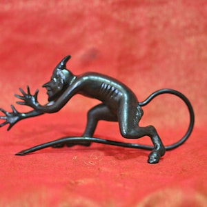 Puede incluir: Figura de bronce oscuro de una criatura parecida a un diablo con cuernos, una larga cola y los brazos extendidos. La figura está en posición agachada, con una larga cola curvada hacia arriba. El fondo es de color rojo liso.