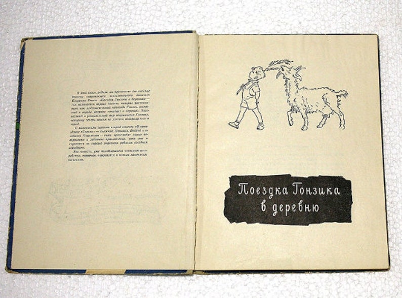 May include: An open book with a black and white illustration of a boy walking a goat. The text on the page reads "Поездка Тонзика в деревню" which translates to "Tonzik's Trip to the Village".