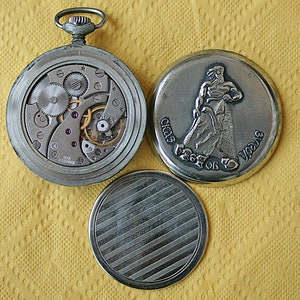 May include: A silver pocket watch with a mechanical movement visible through the open back. The watch has a silver case with a decorative design on the front. The watch is accompanied by a silver case with a design of a person in a traditional costume.