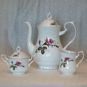 Puede incluir: Un juego de té de porcelana blanca con un diseño floral con rosas rosas y hojas verdes. El juego incluye una tetera, una jarra de crema y un azucarero.