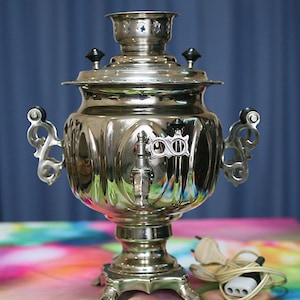 Puede incluir: Un samovar eléctrico plateado con un cable y un enchufe negros. El samovar tiene un cuerpo redondo con un pico y un asa a cada lado. La parte superior del samovar tiene una tapa con un pomo y una pequeña abertura para el vapor.