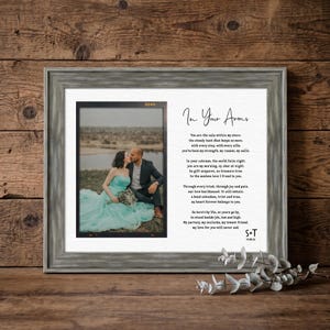 May include: A framed photo print featuring a couple embracing, with a poem titled "In Your Arms" on the right. The frame is gray, and the background is a wooden surface. The photo shows a woman in a teal dress and a man in a suit.