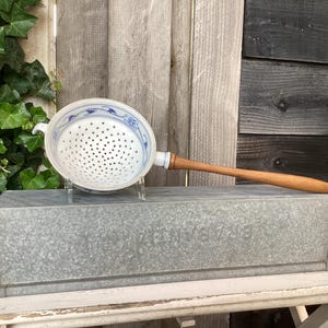 Puede incluir: Un colador de cerámica blanca con detalles florales azules y un mango de madera descansa sobre un recipiente de metal galvanizado. El colador tiene muchos agujeros pequeños para drenar líquidos. El mango es de color marrón claro.
