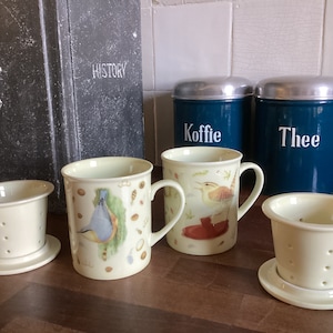 Op de afbeelding: Vier keramische theekopjes met vogelillustraties en een bijpassende theezakjeshouder. De kopjes zijn wit met een crèmekleurige glazuur. De theekopjes hebben handvatten en zijn versierd met vogels en andere natuur geïnspireerde ontwerpen. De theezakjeshouder is wit met een crèmekleurige glazuur en heeft een ronde basis.